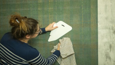 A person holding a rag and using a spatula to smooth wallpaper at home. The wallpaper is green with golden lines.
