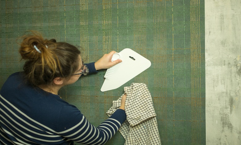 A person holding a rag and using a spatula to smooth wallpaper at home. The wallpaper is green with golden lines.