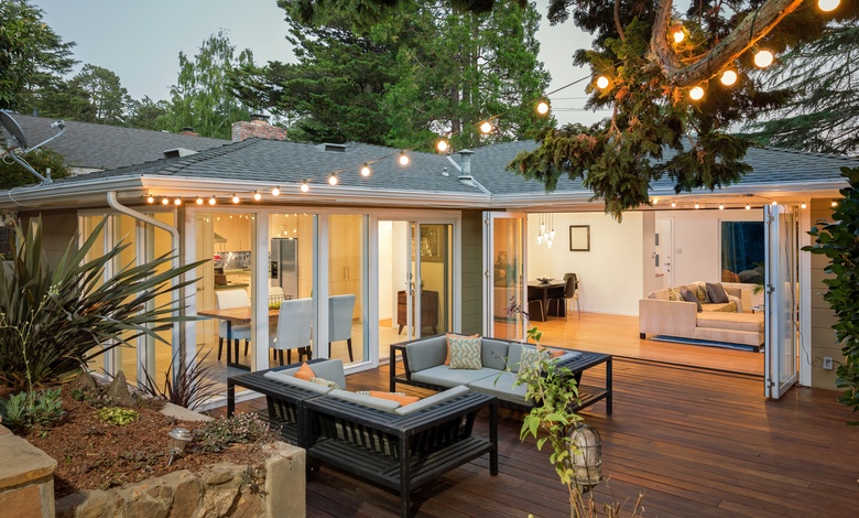 The patio of a modern home with hardwood floors, two loveseat sofas, string lights, and open foldable doors to the house.