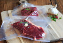Two sealed sous vide bags that contain raw steaks garnished with garlic, herbs, and cracked pepper on a wooden cutting board.