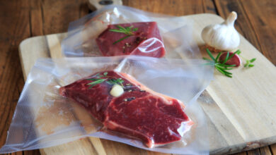 Two sealed sous vide bags that contain raw steaks garnished with garlic, herbs, and cracked pepper on a wooden cutting board.