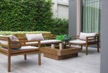 A set of wooden outdoor furniture sits outside a home's window next to several bushes and shrubs growing on the property.