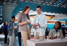 A man and woman standing outside a trade show booth at a conference while another woman sits beside them.