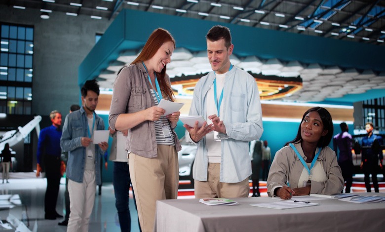 A man and woman standing outside a trade show booth at a conference while another woman sits beside them.