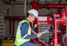 A worker in safety gear inspects a red industrial machine with pipes and valves. He holds a clipboard in one hand.