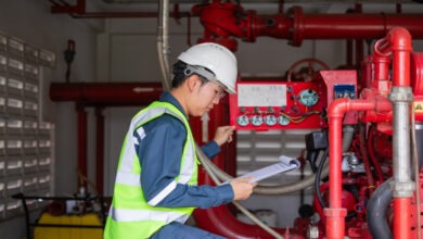 A worker in safety gear inspects a red industrial machine with pipes and valves. He holds a clipboard in one hand.
