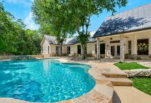 A pool sits on the edge of a patio in a home's backyard, with sparkling blue water. A tree stands near it.