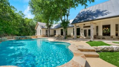A pool sits on the edge of a patio in a home's backyard, with sparkling blue water. A tree stands near it.