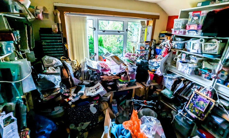 A cluttered room stacked floor to ceiling with hoarded items strewn about and a window letting in light on one end.
