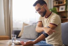A young man wearing glasses sits on a sofa in his home, smiling as he uses a blood pressure monitor.