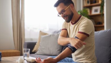 A young man wearing glasses sits on a sofa in his home, smiling as he uses a blood pressure monitor.