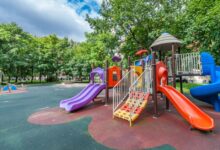A colorful age-inclusive playground with multiple slides, climbing features, and safety surfacing in a tree-lined park setting.