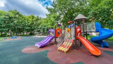 A colorful age-inclusive playground with multiple slides, climbing features, and safety surfacing in a tree-lined park setting.
