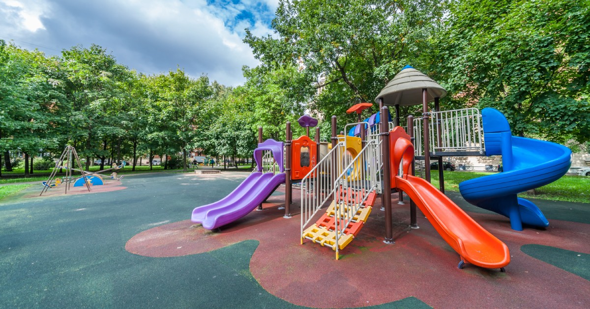 A colorful age-inclusive playground with multiple slides, climbing features, and safety surfacing in a tree-lined park setting.