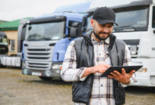 A man wearing a plaid shirt, black vest, and cap holding a tablet while standing in front of semi-trucks in a lot.